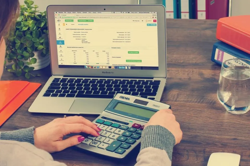 Hands on a calculator beside a laptop showing a spreadsheet